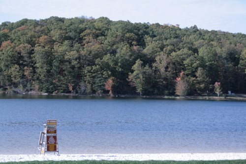 A calm lake surrounded by trees, with a lifeguard stand on a sandy beach in the foreground.