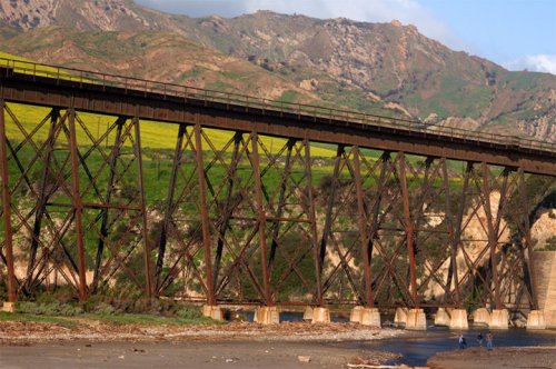 A rusted railway bridge spans a river, surrounded by green hills and mountains under a clear blue sky.