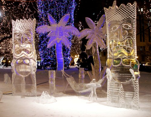 Two large ice sculptures of tiki figures stand among palm trees, illuminated by colorful lights in a winter setting.