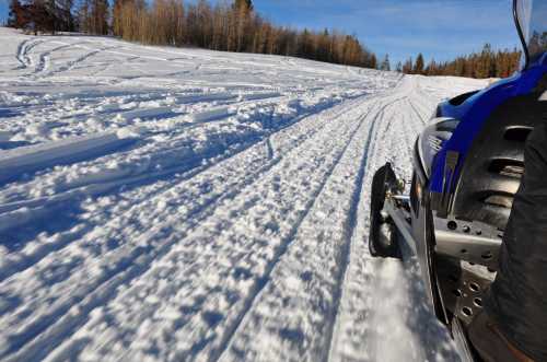 A close-up view of a snowmobile on a snowy trail, with tire tracks in the fresh snow and trees in the background.