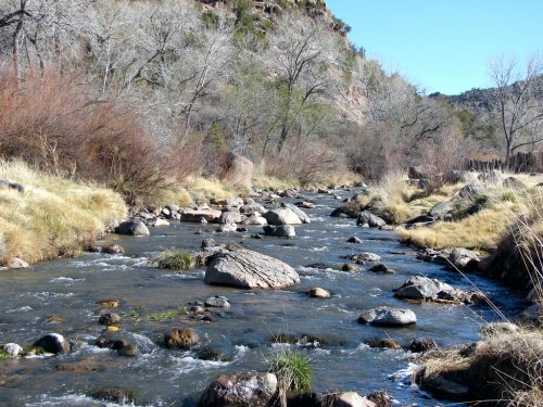 A serene river flows through a rocky landscape, surrounded by trees and dry grass under a clear blue sky.