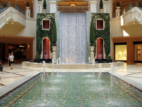 Indoor waterfall cascading down a stone wall, surrounded by greenery and a pool with fountains in a luxurious setting.