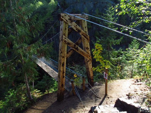 A rustic wooden suspension bridge surrounded by lush green trees and rocky terrain.