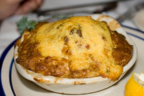A baked dish with a golden, cheesy topping, served in a white bowl on a plate with a lemon wedge.