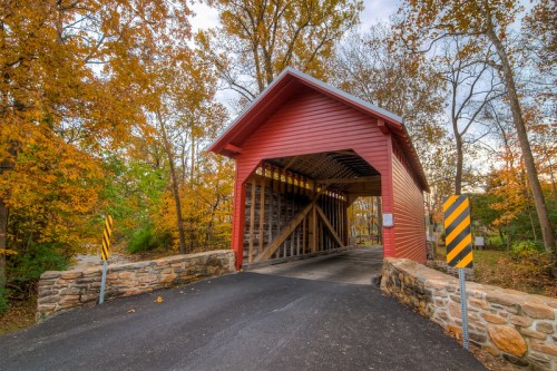A red covered bridge surrounded by autumn trees, with a stone entrance and a paved road leading through it.