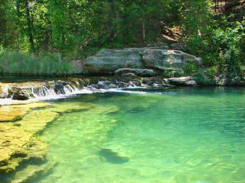 A serene, clear blue-green stream flows over rocks, surrounded by lush greenery and trees.