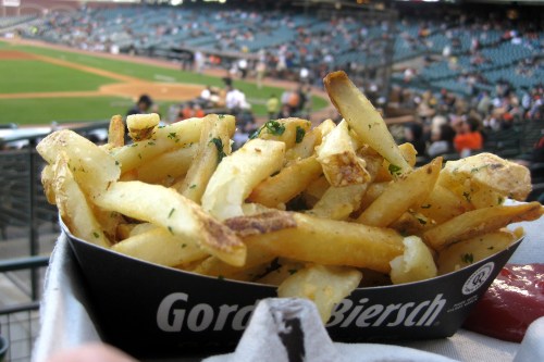 A serving of golden French fries in a black container, with a baseball stadium in the background.