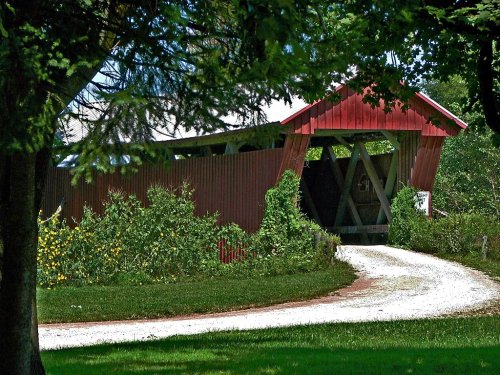 A red covered bridge surrounded by greenery, with a gravel path leading to it under a clear blue sky.