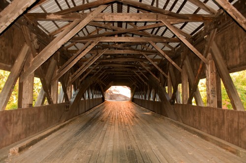 Interior view of a wooden covered bridge, showcasing its rustic beams and a warm glow at the far end.