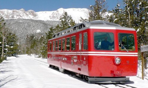 A red train on snowy tracks, surrounded by pine trees and mountains under a clear blue sky.