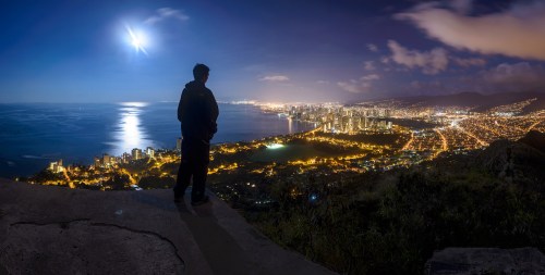 A person stands on a cliff overlooking a city illuminated at night, with a bright moon reflecting on the ocean.