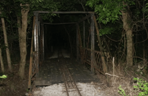 A dark, overgrown bridge with railroad tracks leading into a dense, shadowy forest.