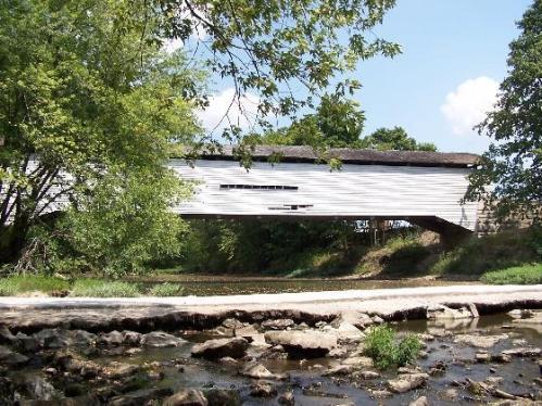A white covered bridge spans a calm stream, surrounded by lush green trees and a clear blue sky.