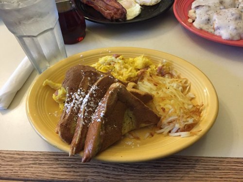 A plate of breakfast featuring French toast, scrambled eggs, hash browns, and a glass of water in the background.