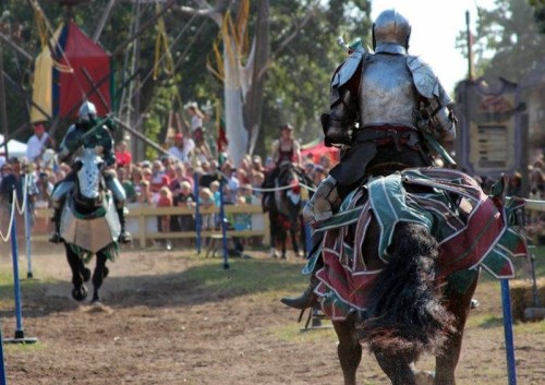 Two armored knights on horseback compete in a jousting event, with a crowd watching in the background.