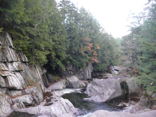 A serene river flows through rocky terrain, surrounded by lush green trees and hints of autumn colors.