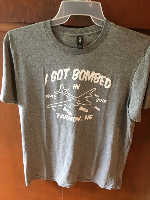 Gray t-shirt hanging on a hanger, featuring the text "I GOT BOMBED IN TARNOV, NE 1943 2018" with airplane graphics.