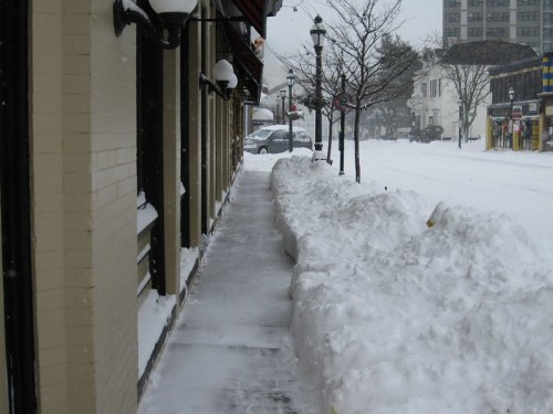 Snow-covered sidewalk beside a building, with large snowdrifts and a quiet street in a winter scene.