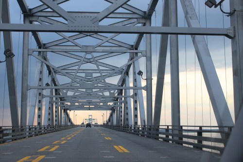 View of a metal bridge structure with a vehicle driving towards the horizon under a cloudy sky.