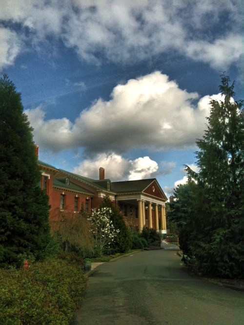 A grand brick building with columns, surrounded by lush greenery and a blue sky with fluffy clouds.