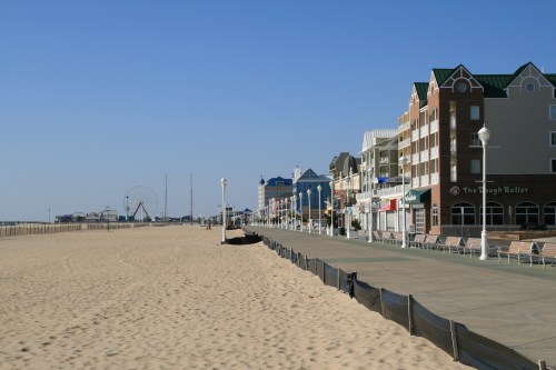 A sandy beach with a boardwalk lined by colorful buildings and a distant ferris wheel under a clear blue sky.