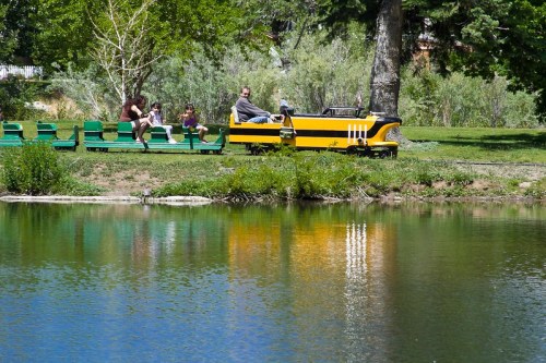 A man sits in a yellow pedal boat on a pond, while two children and a woman relax on a green bench nearby.