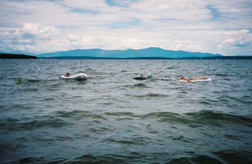 Three people relax on floating devices in a calm lake, with mountains and cloudy skies in the background.