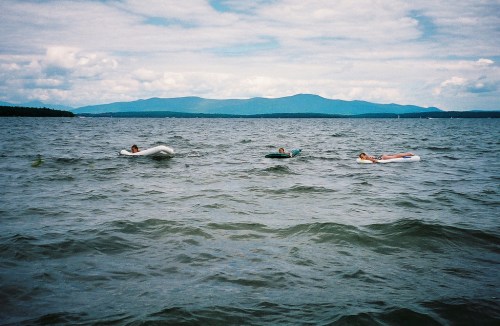 Three people relax on floating devices in a calm lake, with mountains and cloudy skies in the background.