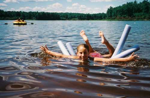 A girl floats in a lake with pool noodles, smiling and splashing, while another person kayaks in the background.