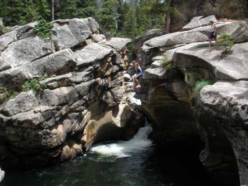 Two people near a rocky gorge with a flowing river, one jumping between rocks and the other standing on a ledge.