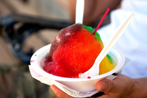 A colorful bowl of shaved ice with red, yellow, and green layers, topped with a straw and two spoons.