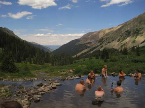 A group of people relaxes in a natural hot spring surrounded by mountains and lush greenery under a blue sky.