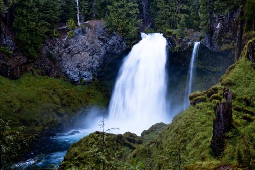 A majestic waterfall cascades over rocky cliffs, surrounded by lush green vegetation and mist rising from the water.