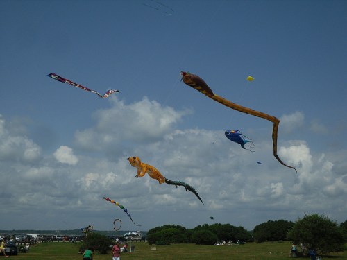Colorful kites of various shapes soar in a blue sky with fluffy clouds over a grassy field.