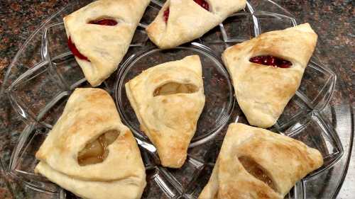 A plate of six golden pastries with fruit fillings, featuring visible openings filled with red and yellow jam.