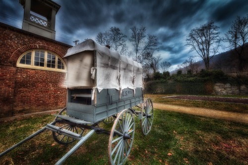 A vintage wagon with a white cover sits on grass, surrounded by trees and a cloudy sky.