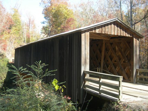 A wooden covered bridge surrounded by autumn foliage and greenery, with a small walkway leading to it.