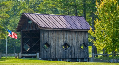 A rustic wooden covered bridge with a red roof, surrounded by trees and an American flag nearby.