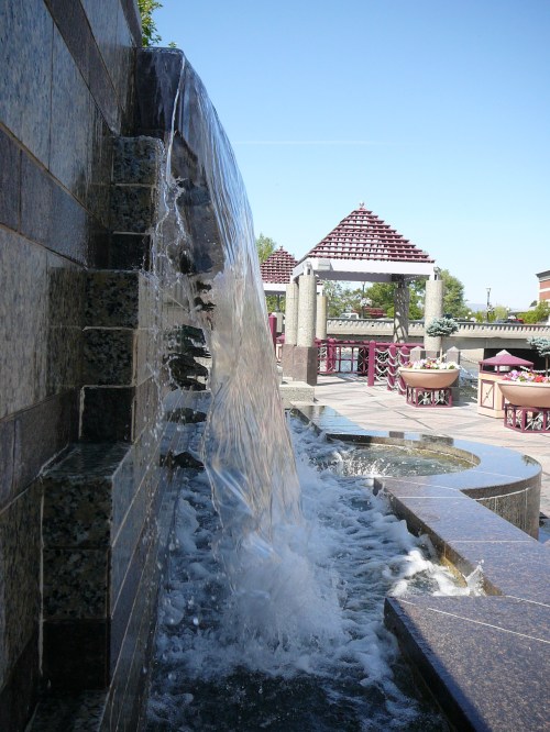 A cascading water feature flows over stone, surrounded by a landscaped area with seating and decorative structures.