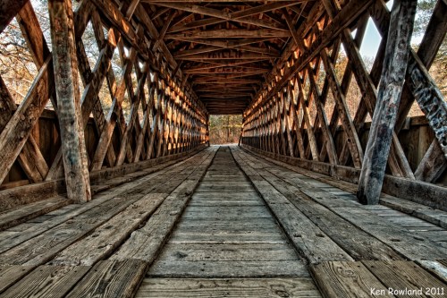 A wooden covered bridge with a lattice design, featuring a weathered wooden floor and warm sunlight filtering through.
