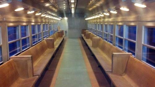 Interior of a train car with wooden benches and large windows, featuring a long, empty aisle.