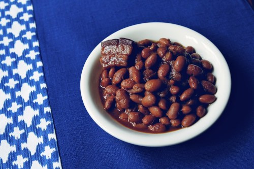 A bowl of cooked pinto beans with a piece of meat, placed on a blue tablecloth with a patterned edge.