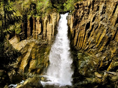 A cascading waterfall flows over rocky cliffs, surrounded by lush greenery and natural rock formations.