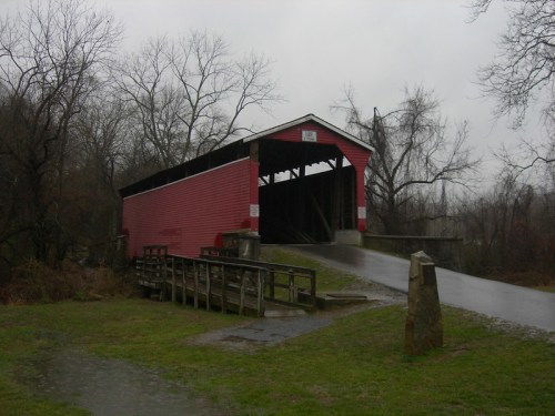 A red covered bridge stands over a wet road, surrounded by bare trees and a gray, overcast sky.