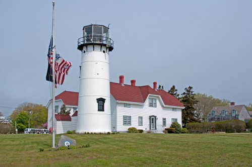 A white lighthouse with a red roof, surrounded by green grass and flags, under a cloudy sky.