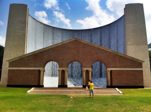 A large brick structure with arches in front of a cascading waterfall, set against a blue sky. Two people are taking photos.