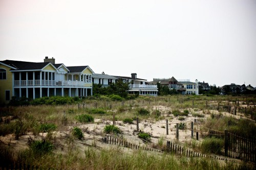 Coastal scene featuring beach houses along a sandy shore with grass and wooden fences in the foreground.