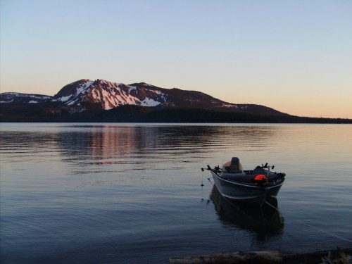 A calm lake at sunset with a boat in the foreground and snow-capped mountains in the background.