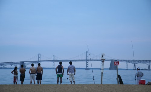 A group of people fishing on a beach, with a bridge visible in the background under a cloudy sky.