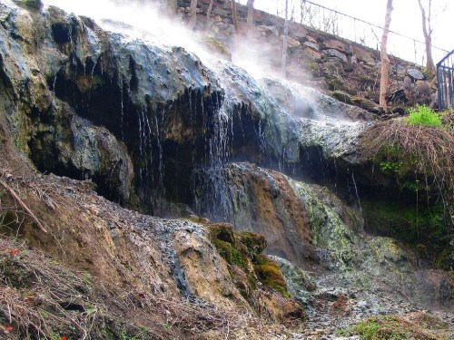 A rocky waterfall with steam rising, surrounded by greenery and a stone path in the background.
